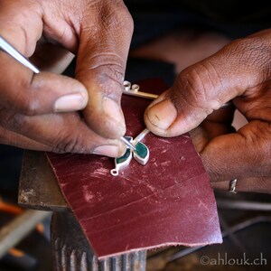 May include: A person's hands are working on a silver pendant with green gemstones. The pendant is being polished on a red surface.