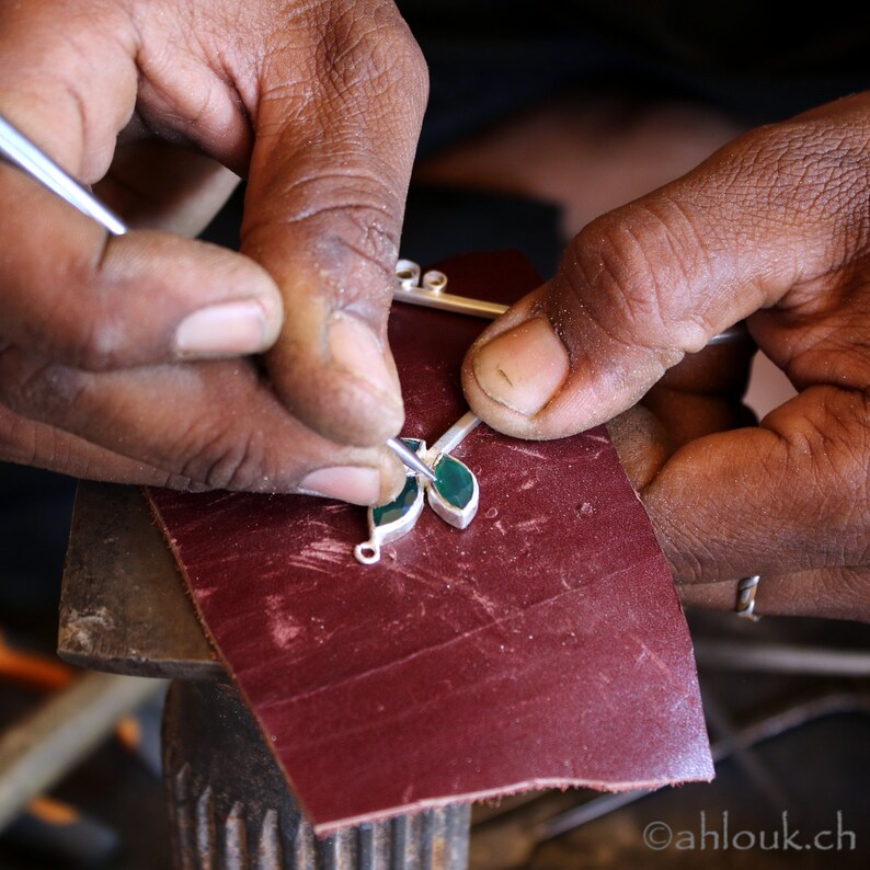 May include: A person's hands are working on a silver pendant with green gemstones. The pendant is being polished on a red surface.