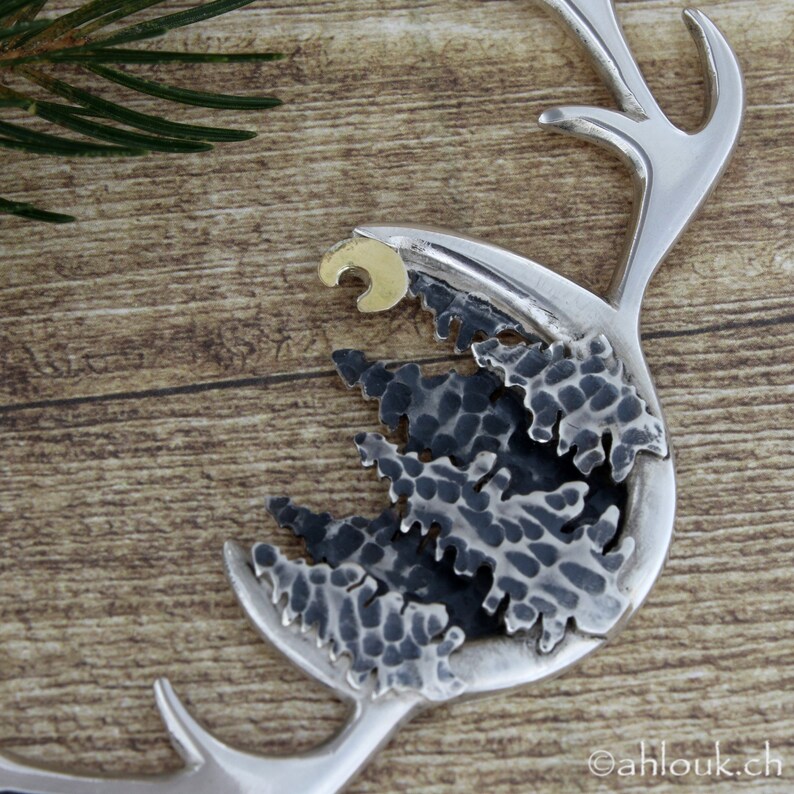 May include: A silver antler pendant with a hammered metal design featuring a crescent moon and a cluster of stylized pine trees. The pendant is on a wooden surface.