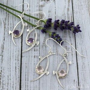 Pu&ograve; includere: Un paio di orecchini d'argento con un design delicato e astratto. Ogni orecchino presenta una pietra preziosa viola o rosa incastonata in una montatura d'argento. Gli orecchini sono fotografati su uno sfondo di legno bianco con rametti di lavanda.