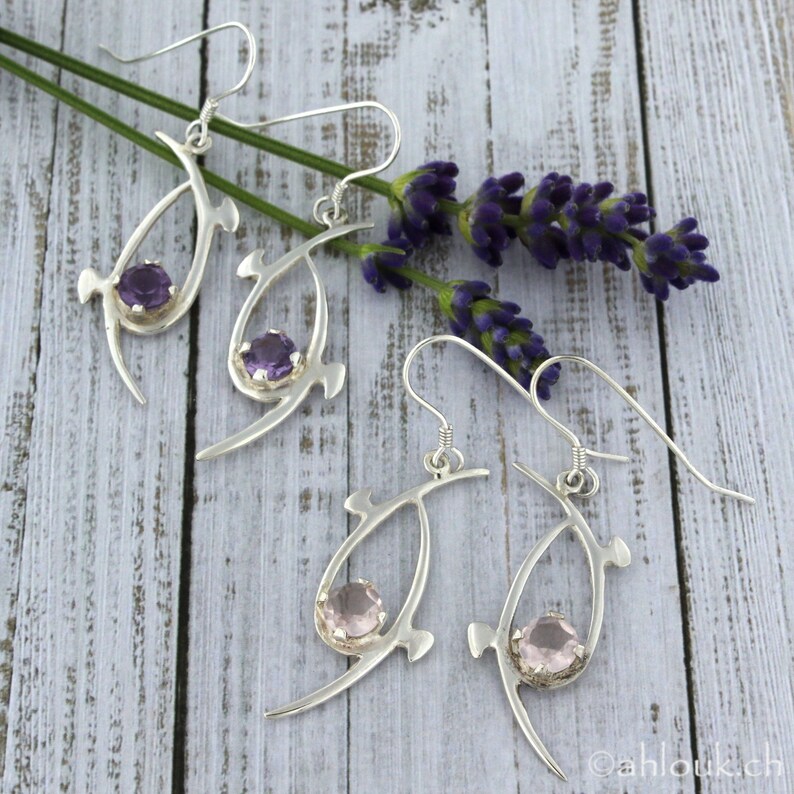 Pu&ograve; includere: Un paio di orecchini d'argento con un design delicato e astratto. Ogni orecchino presenta una pietra preziosa viola o rosa incastonata in una montatura d'argento. Gli orecchini sono fotografati su uno sfondo di legno bianco con rametti di lavanda.