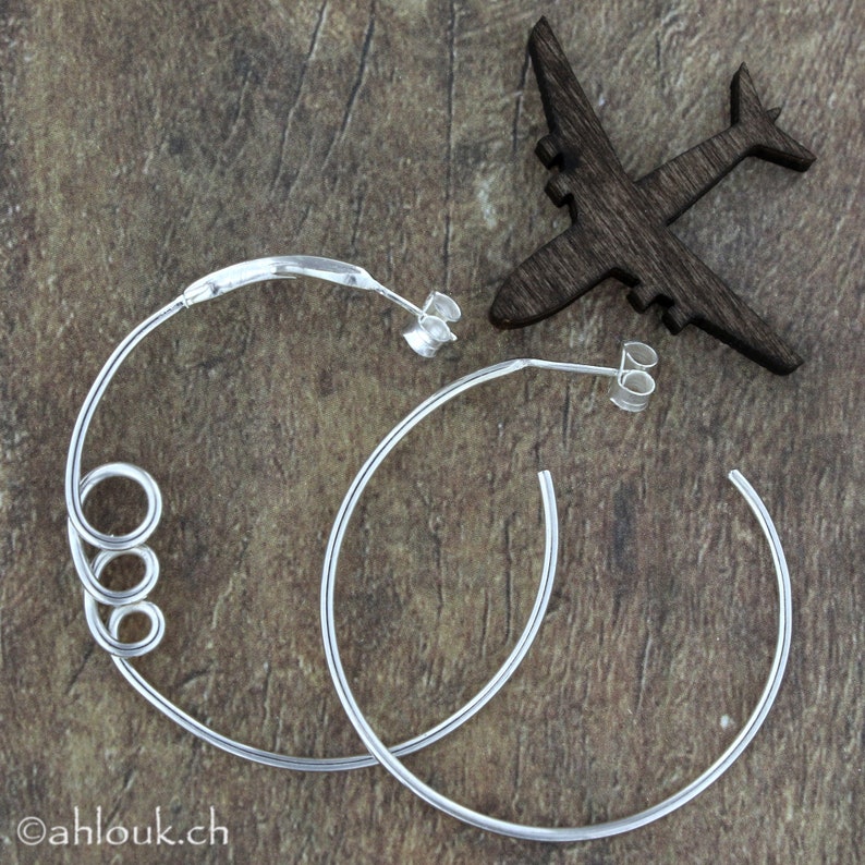 May include: A pair of silver hoop earrings. One earring has a twisted design, while the other is a simple hoop. The earrings are on a wooden surface.