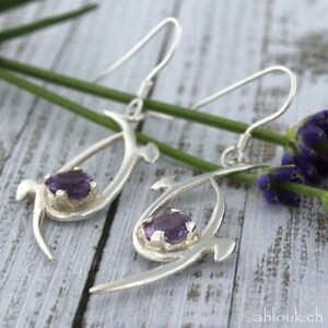 Pu&ograve; includere: Un paio di orecchini d'argento con un design delicato e astratto. Ogni orecchino presenta una pietra preziosa di ametista viola incastonata in una montatura d'argento. Gli orecchini sono sospesi da ganci d'argento.