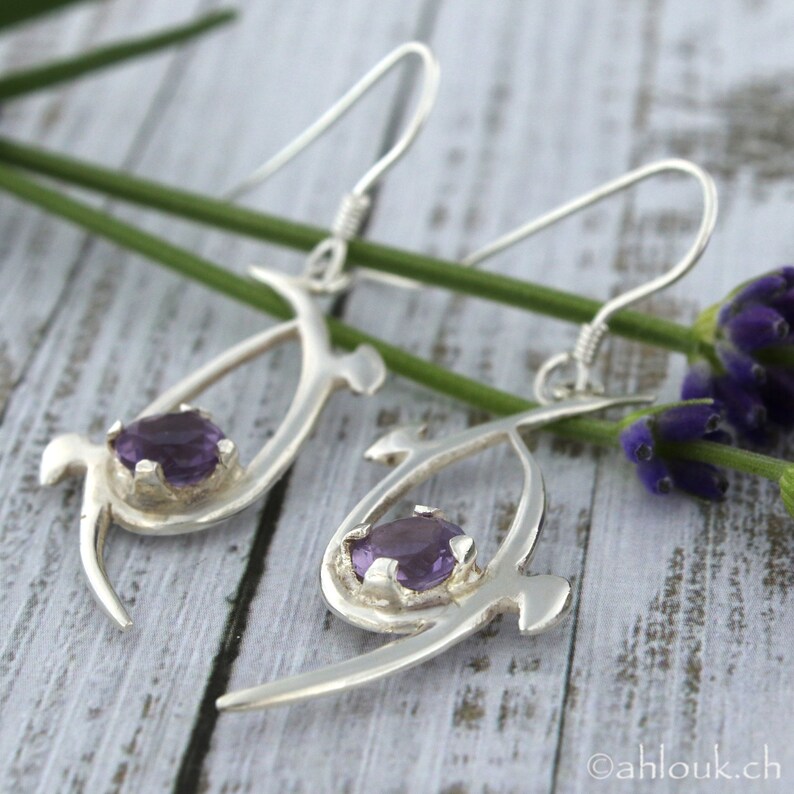 Pu&ograve; includere: Un paio di orecchini d'argento con un design delicato e astratto. Ogni orecchino presenta una pietra preziosa di ametista viola incastonata in una montatura d'argento. Gli orecchini sono sospesi da ganci d'argento.