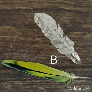 May include: A silver feather ring with a detailed design. The ring is on a wooden surface with a green and yellow feather.