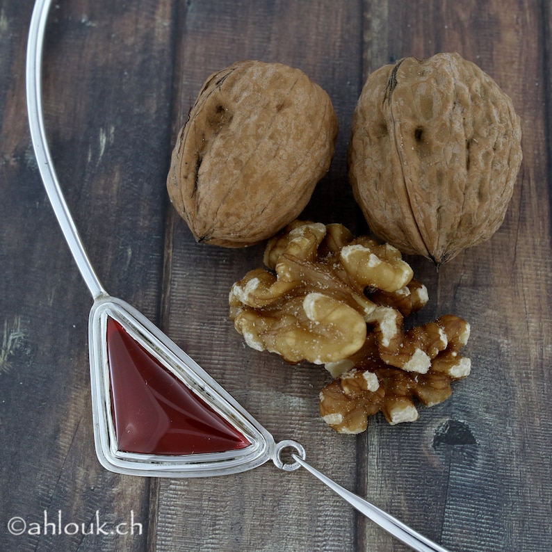 Peut inclure: Un collier en argent avec un pendentif triangulaire orn&eacute; d'une pierre pr&eacute;cieuse rouge. Le collier est pr&eacute;sent&eacute; sur une surface en bois avec trois noix.