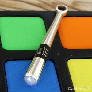 May include: A silver and black metal pipe with a clear gemstone at the end. The pipe is resting on a colorful palette with green, orange, blue, and yellow squares.