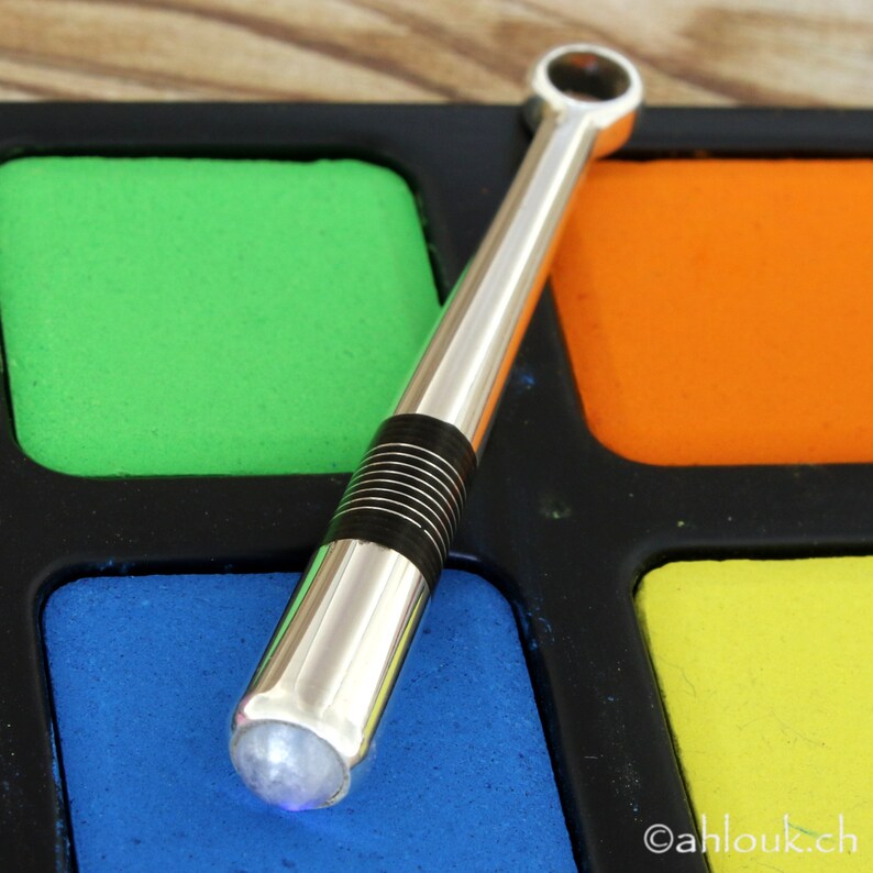 May include: A silver and black metal pipe with a clear gemstone at the end. The pipe is resting on a colorful palette with green, orange, blue, and yellow squares.