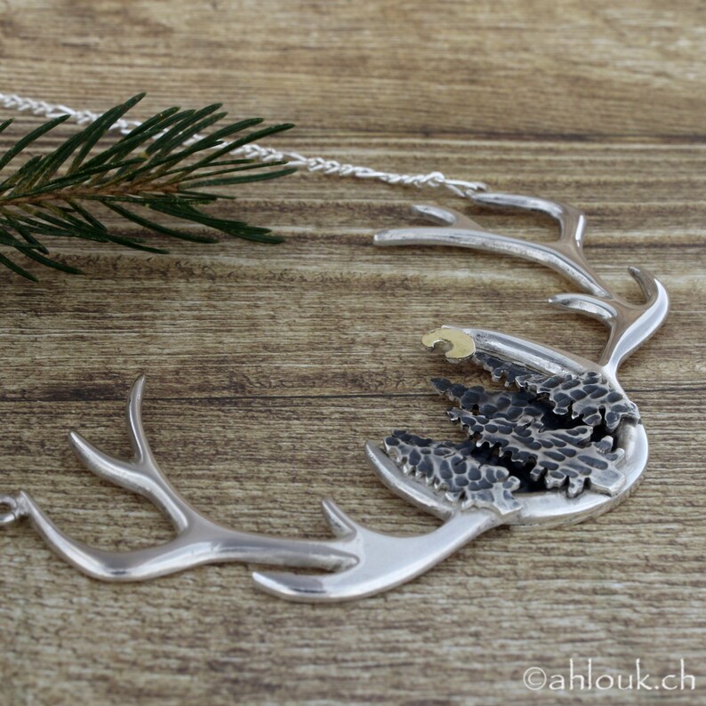 May include: A silver necklace with a deer antler design. The antlers are textured and have a small gold accent. The antlers are holding a cluster of black and silver textured trees.