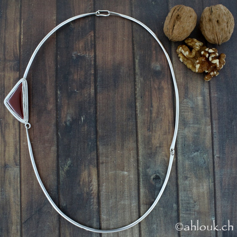 Peut inclure: Un collier ras de cou argent&eacute; avec un pendentif en forme de triangle en pierre de cornaline. Le collier est sur un fond en bois.