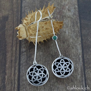 May include: A pair of silver dangle earrings with a black and silver geometric design and a green gemstone accent. The earrings are hanging from a brown, spiky, dried plant.