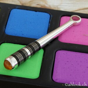 May include: A silver baseball bat pendant with a black and white striped design and a clear amber stone at the end. The pendant is on a colorful background.