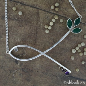 May include: Silver necklace with a stylized fish design and a small green leaf pendant. The pendant has three green leaves and a small clear stone in the center. The necklace is on a wooden surface with white peppercorns.