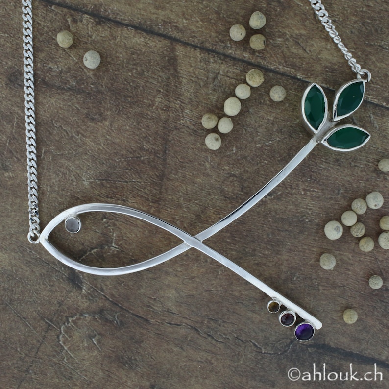 May include: Silver necklace with a stylized fish design and a small green leaf pendant. The pendant has three green leaves and a small clear stone in the center. The necklace is on a wooden surface with white peppercorns.