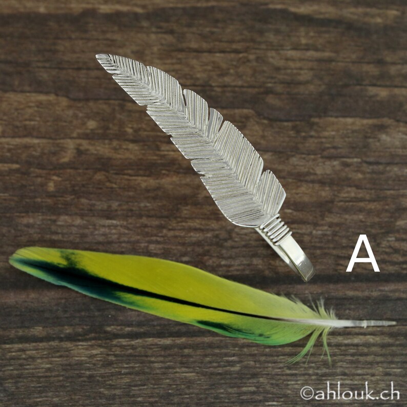 May include: A silver feather ring with a detailed design. The ring is on a wooden surface with a green and yellow feather.