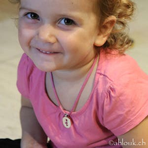 May include: A young girl wearing a pink shirt with a wooden button necklace. The button is a light brown color and has a simple design.
