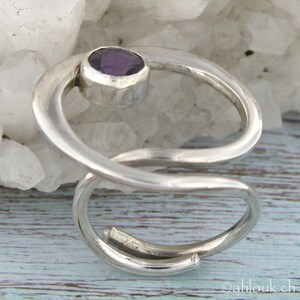 May include: A silver ring with a purple amethyst gemstone set in a curved band. The ring is designed with a twisted, open-ended band.