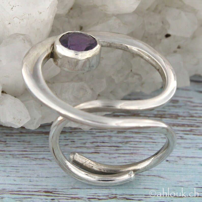 May include: A silver ring with a purple amethyst gemstone set in a curved band. The ring is designed with a twisted, open-ended band.