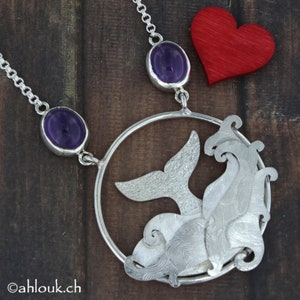 May include: A silver necklace with a whale tail pendant and two amethyst stones. The pendant is in the shape of a whale tail with waves around it. The necklace is on a wooden background.