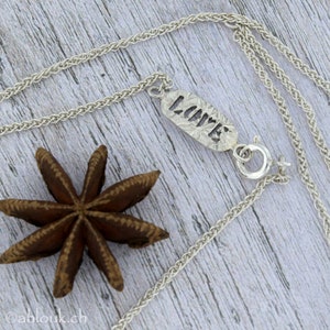 May include: A silver chain necklace with a pendant that says "LOVE" in a textured, cutout font. The pendant is attached to the chain with a silver clasp.