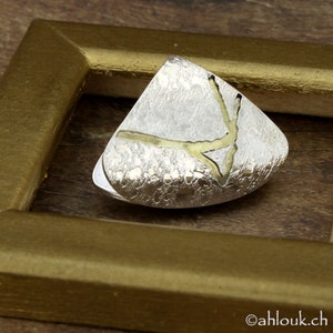 May include: A silver-toned, triangular-shaped brooch with a textured surface and gold-colored accents. The brooch is set within a gold-colored frame, showcasing a unique, abstract design. The brooch is a piece of jewelry.