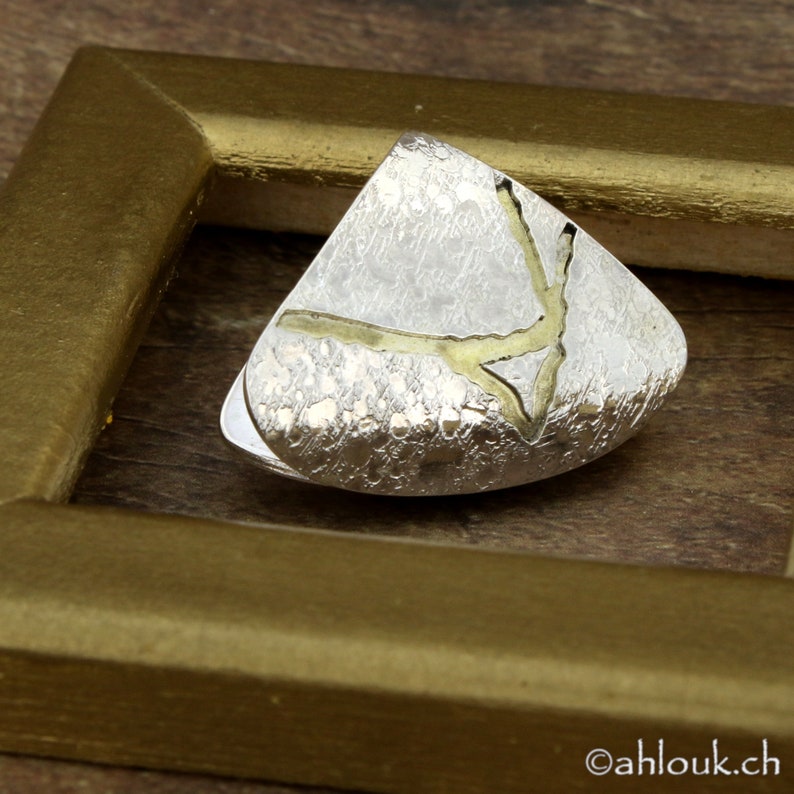 May include: A silver-toned, triangular-shaped brooch with a textured surface and gold-colored accents. The brooch is set within a gold-colored frame, showcasing a unique, abstract design. The brooch is a piece of jewelry.
