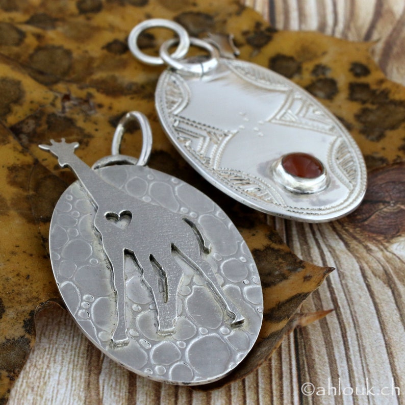 May include: Two silver pendants. One oval pendant features a giraffe silhouette with a heart cutout. The other oval pendant has an engraved design and a small, round, reddish-brown stone. Both pendants have a loop for a chain.