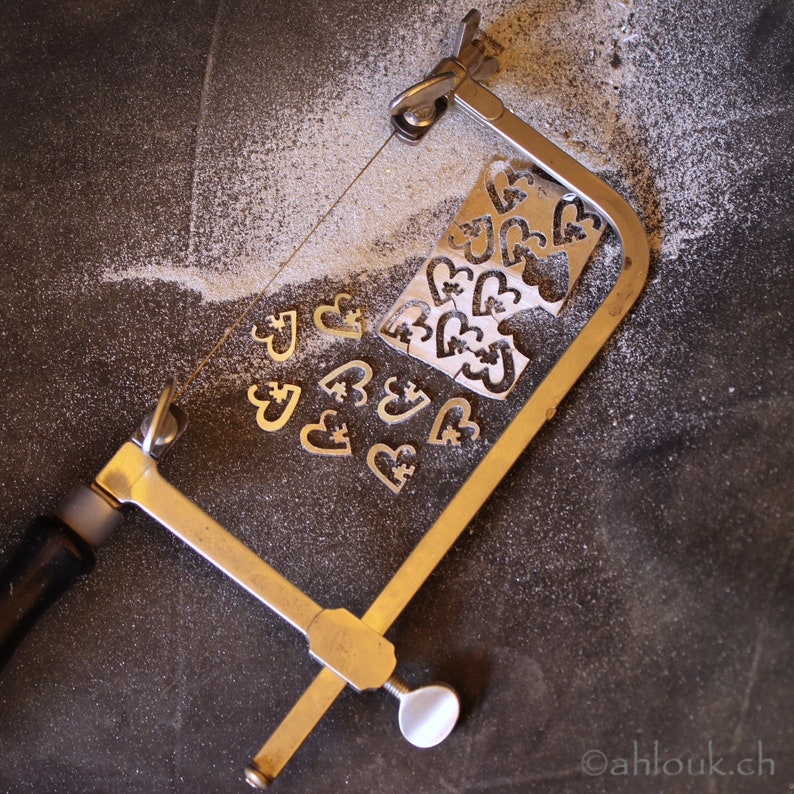 May include: A silver metal saw with a wooden handle is used to cut out small heart shapes from a sheet of metal. The heart shapes are scattered around the saw.