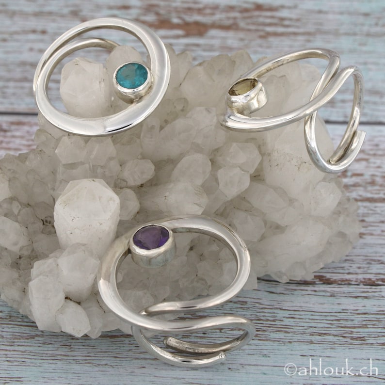 May include: Three silver rings with different colored gemstones. The rings are made of twisted silver and feature a single round gemstone in the center. The gemstones are turquoise, yellow, and purple.