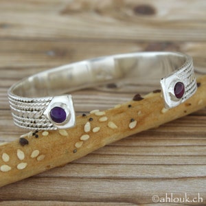 Puede incluir: Pulsera de plata con dos piedras redondas, una morada y una roja. La pulsera est&aacute; sobre una superficie de madera.