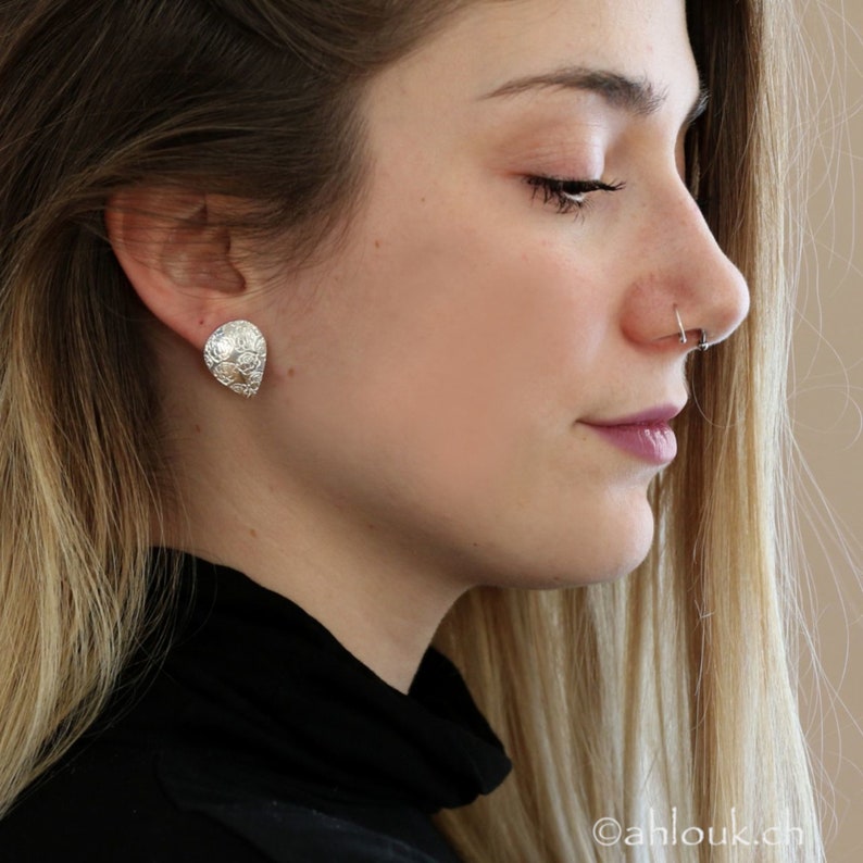 May include: A pair of silver teardrop-shaped earrings with a textured surface. The earrings are worn by a person with blonde hair.