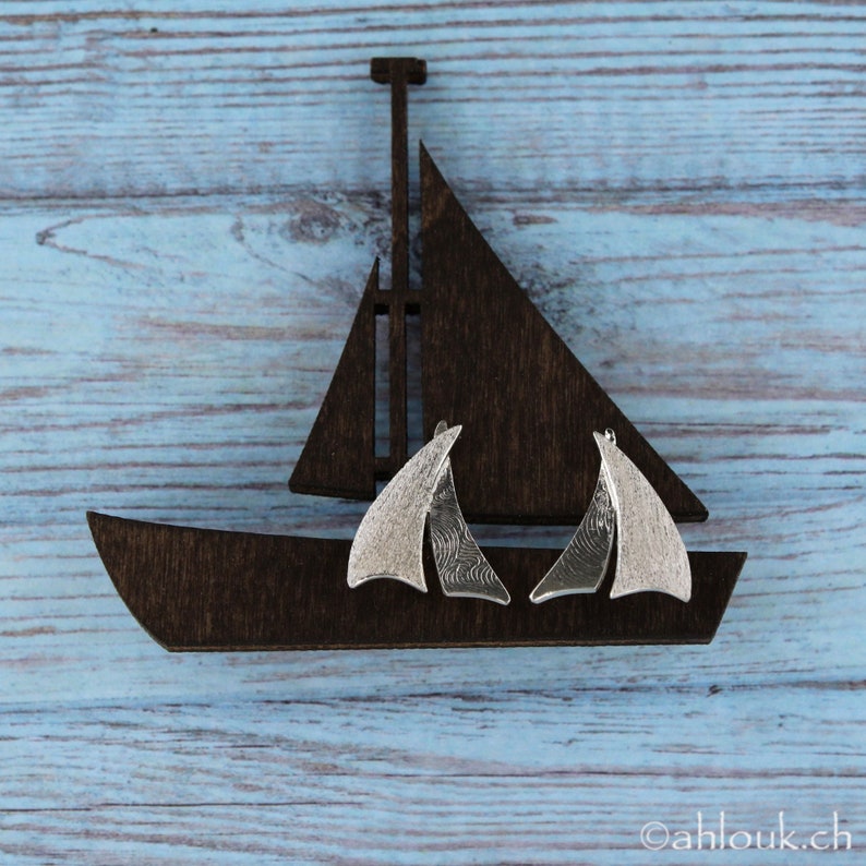 K&ouml;nnte beinhalten: Ein Paar silberne Segelschiff-Ohrringe mit strukturierter Oberfl&auml;che, pr&auml;sentiert auf einem h&ouml;lzernen Segelschiff-Ausschnitt vor einem blauen Holz-Hintergrund.