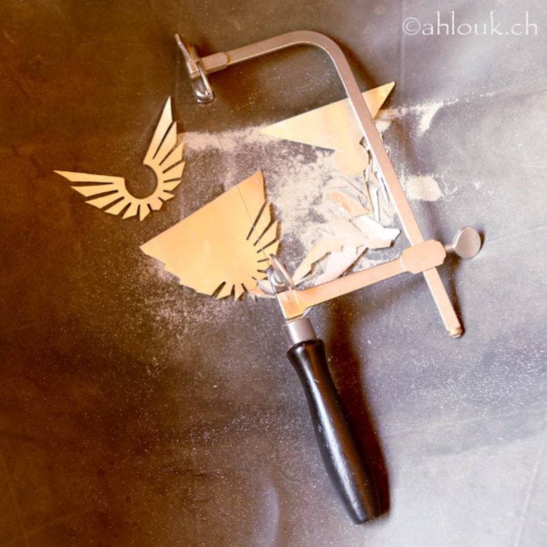 May include: A silver hand saw with a black handle is used to cut a gold metal wing shape. The saw is on a gray surface with sawdust.