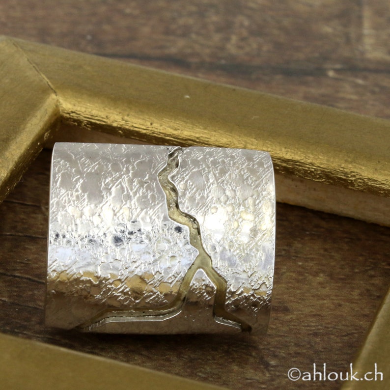 May include: A silver-colored ring with a textured surface and a gold-colored, abstract design. The ring is rectangular and sits within a gold-colored frame. The ring's surface has a hammered appearance. The website address "ahlouk.ch" is visible in the bottom right corner.