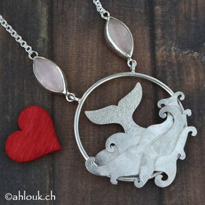 May include: A silver necklace with a whale tail pendant and a wave design. The pendant is surrounded by a circle and hangs from a chain with two pink oval stones.