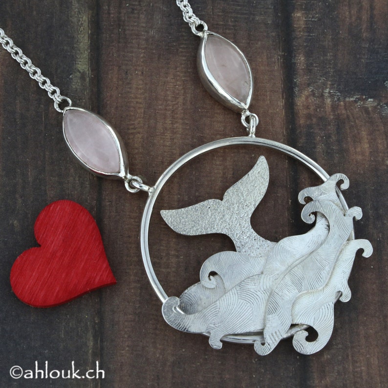 May include: A silver necklace with a whale tail pendant and a wave design. The pendant is surrounded by a circle and hangs from a chain with two pink oval stones.