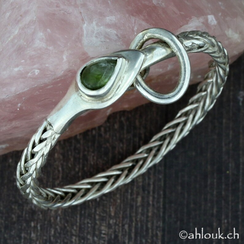 May include: A silver bracelet with a braided chain design and a green gemstone set in a snake head clasp.
