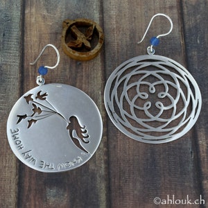 May include: A pair of silver earrings. One earring features a cutout design of a woman holding a bunch of birds with the text "I know the way home". The other earring features a circular cutout design with a floral pattern.