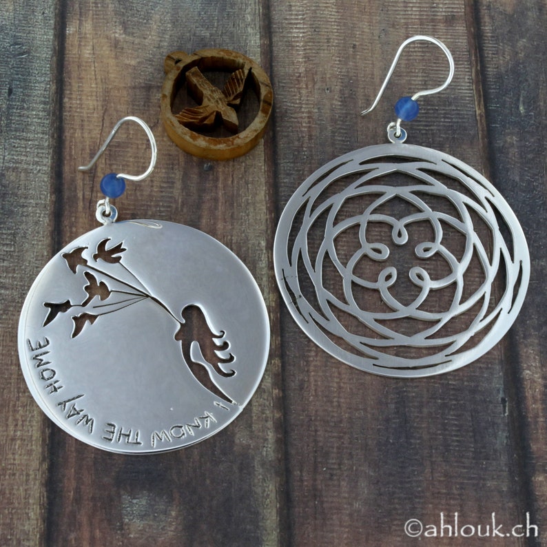 May include: A pair of silver earrings. One earring features a cutout design of a woman holding a bunch of birds with the text "I know the way home". The other earring features a circular cutout design with a floral pattern.