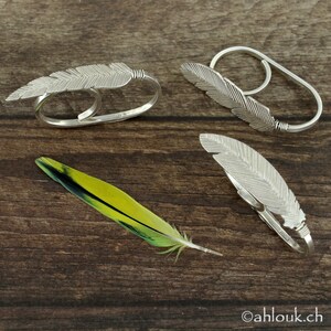 May include: Three silver feather rings on a wooden surface. The rings are designed to fit on two fingers. The rings are made of silver and have a delicate feather design.
