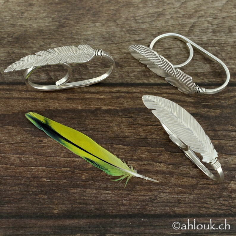 May include: Three silver feather rings on a wooden surface. The rings are designed to fit on two fingers. The rings are made of silver and have a delicate feather design.