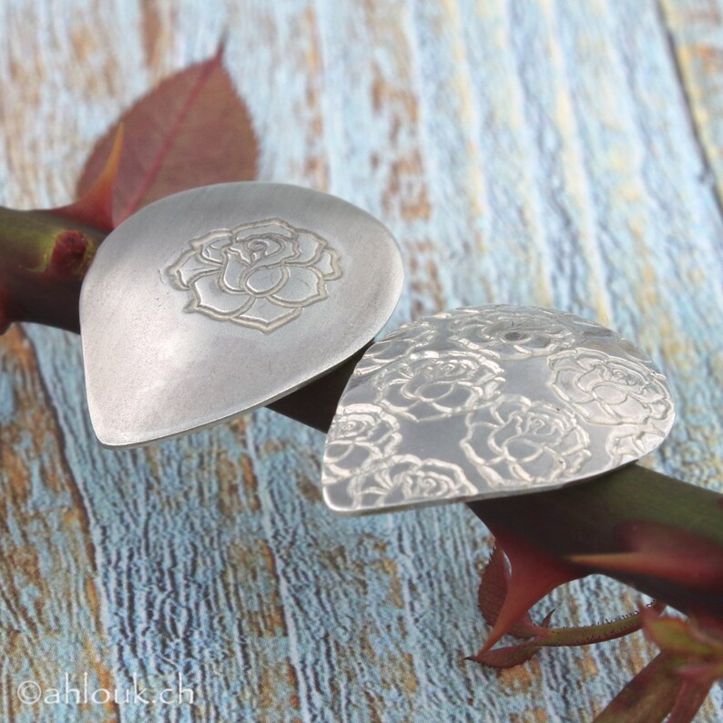May include: Two silver guitar picks with a rose design. The pick on the left has a single rose in the center, while the pick on the right has a pattern of smaller roses.