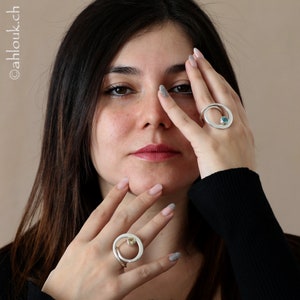 May include: Two silver rings with a circular design. One ring features a turquoise gemstone, while the other ring has a yellow gemstone. The rings are worn on the fingers of a person's hand.