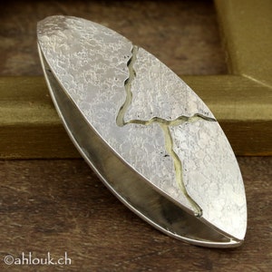 May include: Silver-toned, oval-shaped pendant with a textured surface and a gold-colored inlay resembling a stylized tree or river. The pendant has a polished, smooth edge and a unique, artistic design. The website address "ahlouk.ch" is visible.