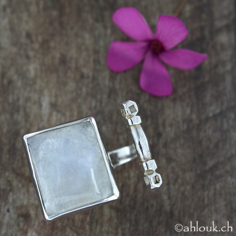 May include: A silver ring featuring a square moonstone and a silver bar design. The ring is displayed on a wooden surface.