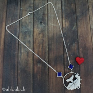 May include: A silver necklace with a pendant featuring two stylized figures and a blue gemstone. The pendant is attached to a silver chain with a lobster clasp. A red heart charm hangs from the chain.