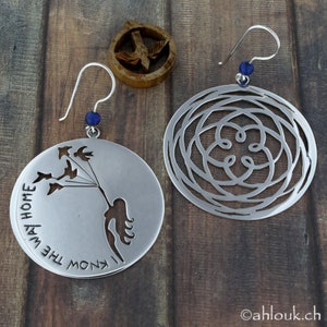 May include: A pair of silver earrings. One earring features a circular design with a cutout of a woman holding birds. The other earring has a circular design with a cutout of a geometric pattern. Both earrings have a blue gemstone on the hook.