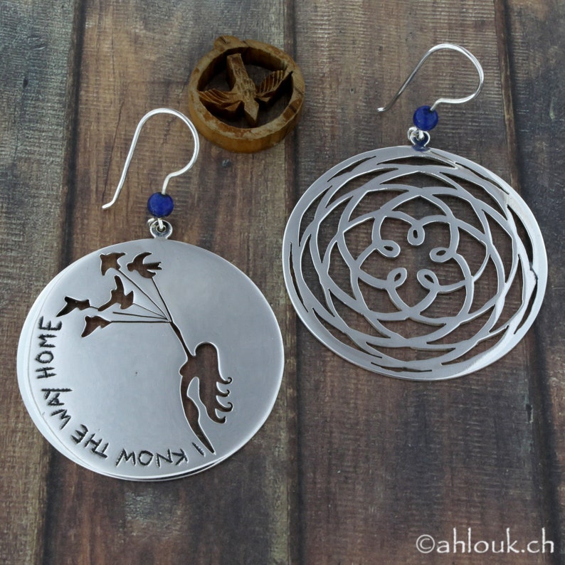 May include: A pair of silver earrings. One earring features a circular design with a cutout of a woman holding birds. The other earring has a circular design with a cutout of a geometric pattern. Both earrings have a blue gemstone on the hook.