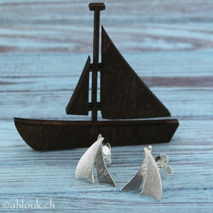 K&ouml;nnte beinhalten: Ein Paar silberne Segelschiff-Ohrringe. Die Ohrringe bestehen aus zwei &uuml;berlappenden Segelformen mit einer strukturierten Oberfl&auml;che. Die Ohrringe werden auf einem Holzsegelschiff pr&auml;sentiert.