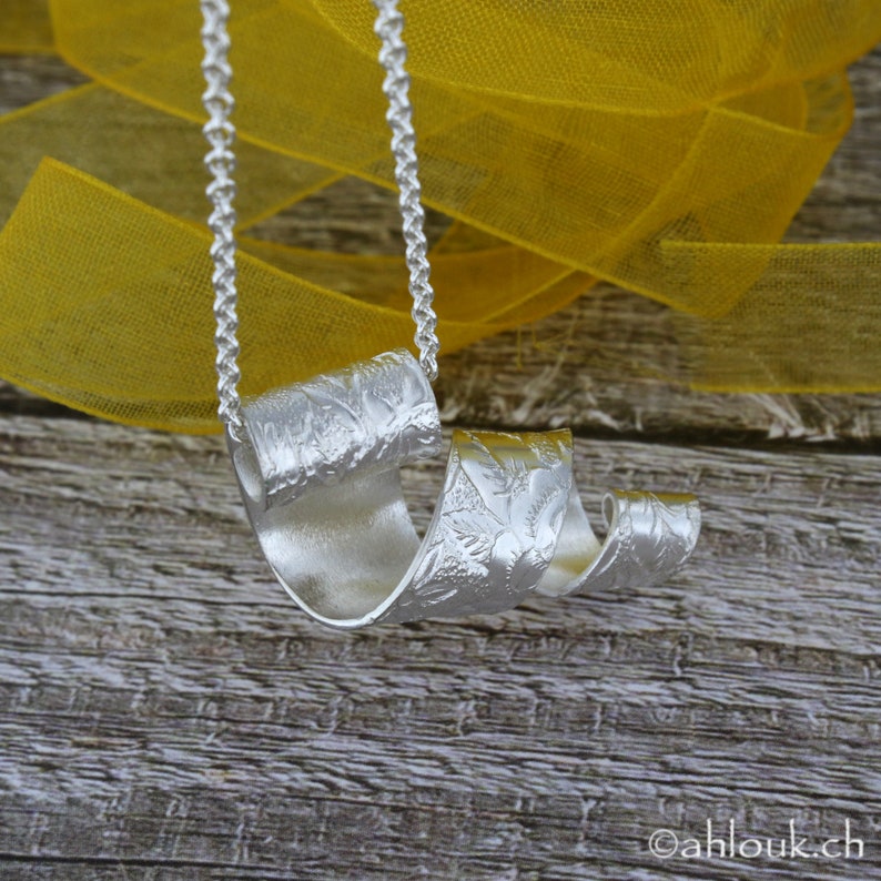 May include: A silver necklace with a unique, textured pendant that is shaped like a swirl. The pendant has a floral design and is hanging from a silver chain.
